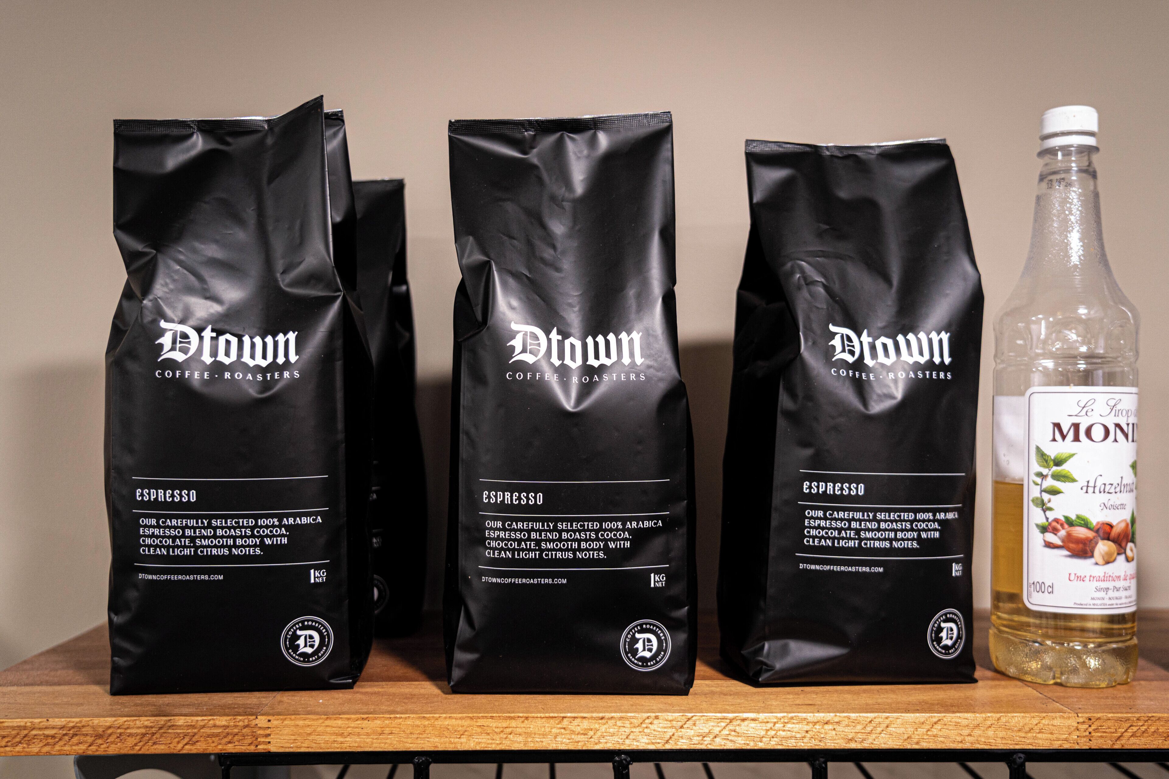 Three bags of Dtown Coffee Roasters espresso and a bottle of Le Tropique Monin hazelnut syrup on a shelf.