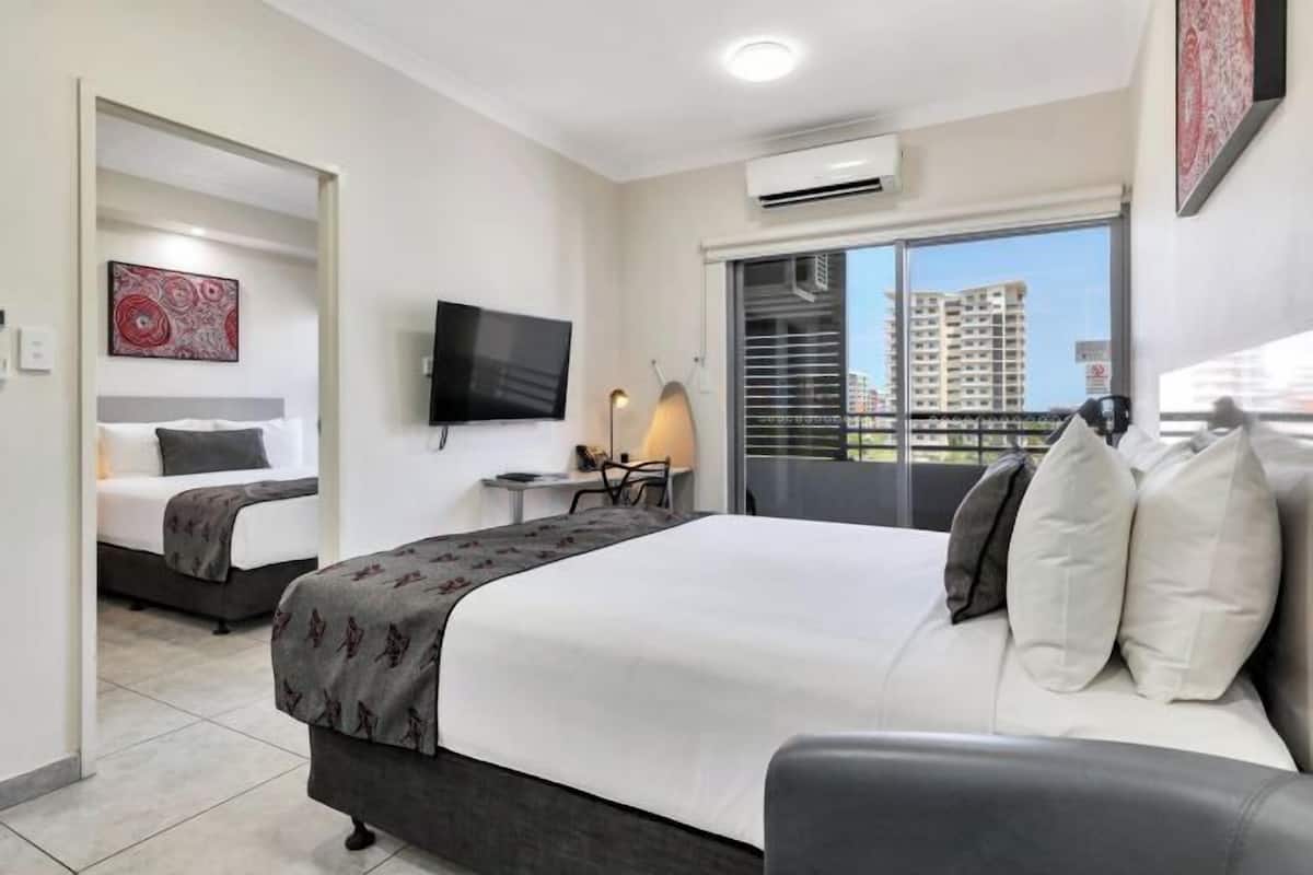 A hotel room with a bed, a desk, a chair, a TV, and a balcony with a view of buildings.