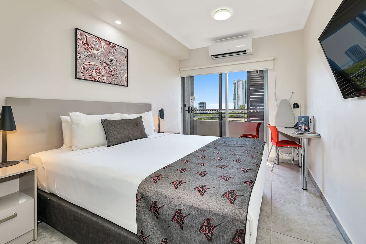 A modern hotel room with a large bed, a desk with a chair, a TV, and a balcony with a city view.