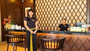 Un área de recepción con dos recepcionistas, un mostrador con varios objetos y dos sillas.