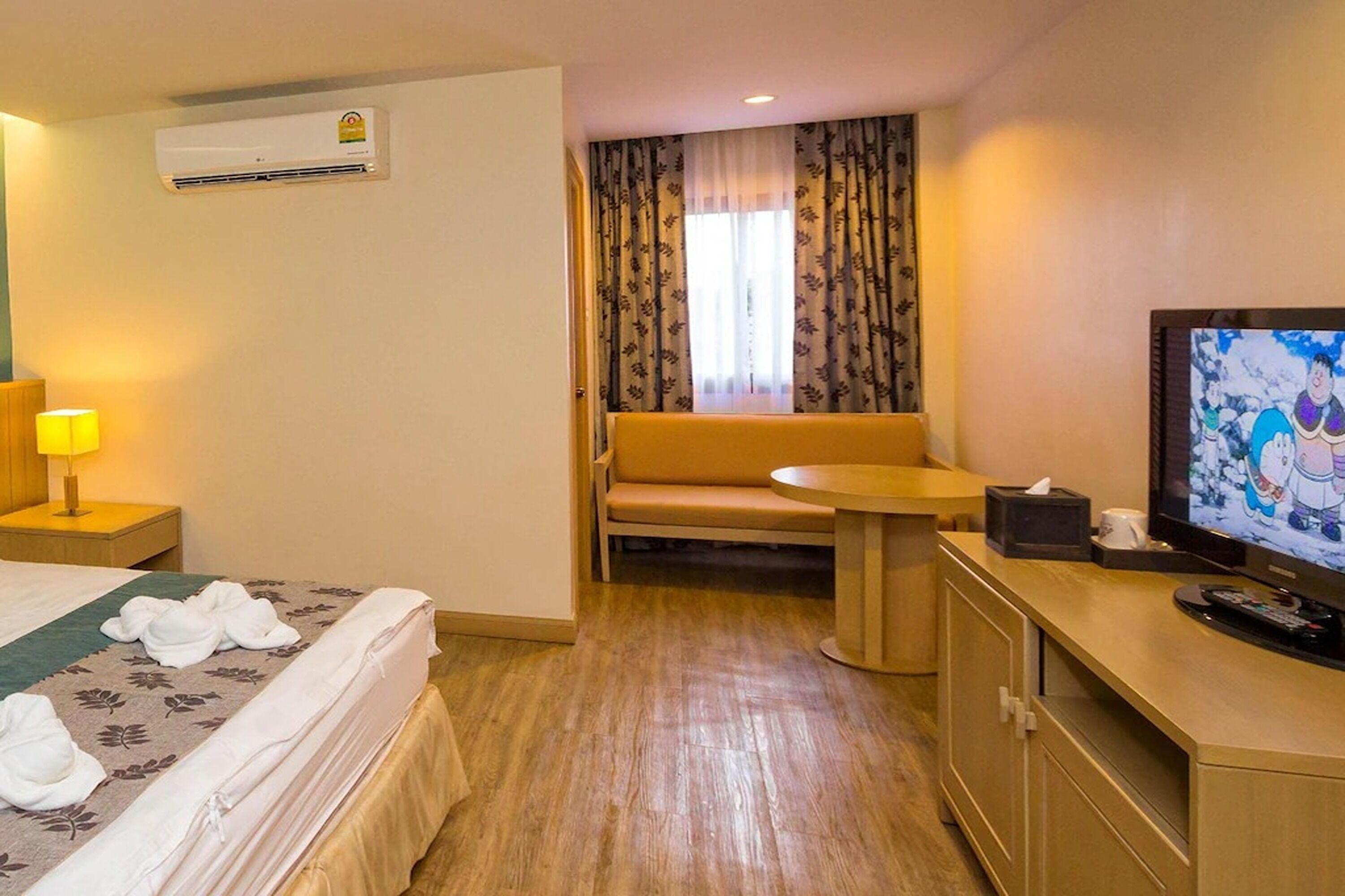 A hotel room with a bed, a sofa, a small table, a television, and an air conditioning unit.