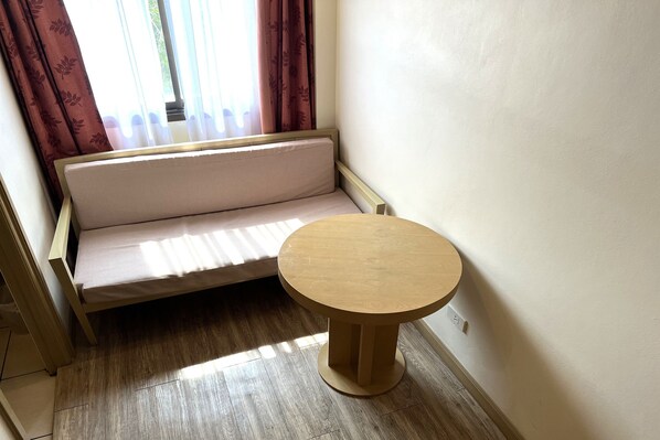 Una habitación con piso de madera, una mesita redonda pequeña, un sofá y una ventana con cortinas.