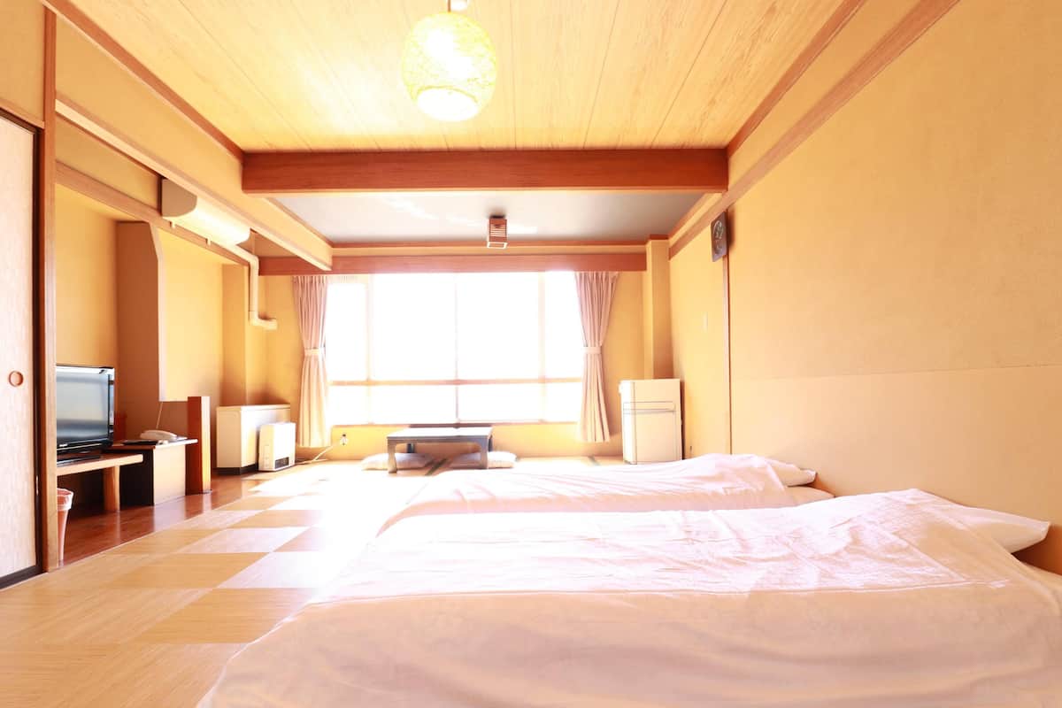 Een hotelkamer met een houten vloer, een bed met wit beddengoed, een bureau met een computer, een televisie en een raam met gordijnen.