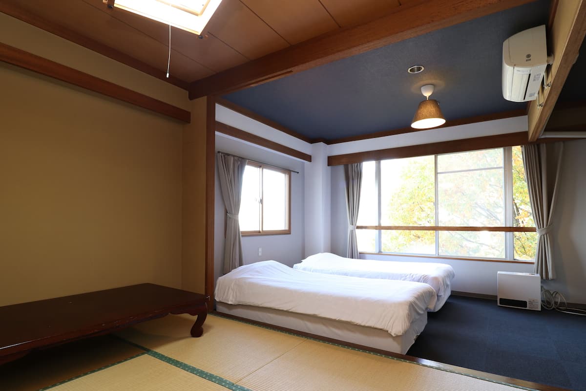 Een kamer met een bed, een houten bankje, tatami vloeren, een plafondventilator en een raam met gordijnen.