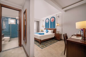 Habitación de hotel con baño, cama con cubrecama azul, mesita de noche, escritorio con lámpara y una silla.
