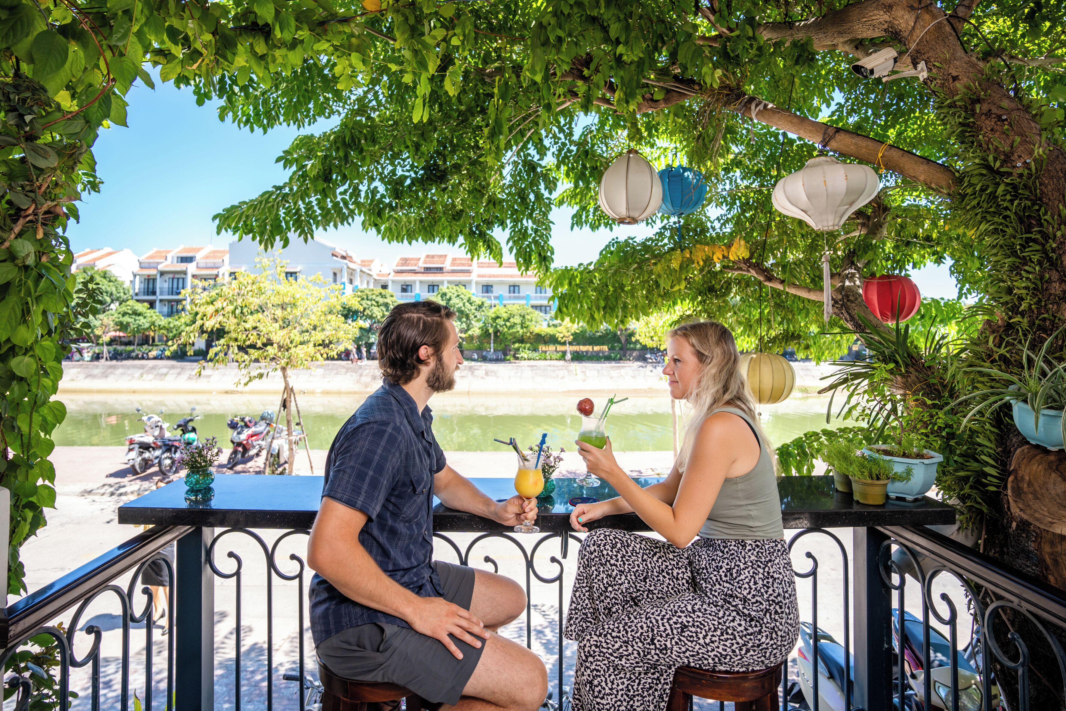 Um terraço com vista para um rio e edifícios, com mesas ao ar livre e plantas em vasos.