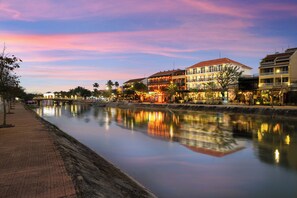 Un hôtel situé au bord de l’eau, dont les bâtiments se reflètent dans les eaux calmes, avec une promenade pavée longeant la rivière et un ciel au coucher du soleil.