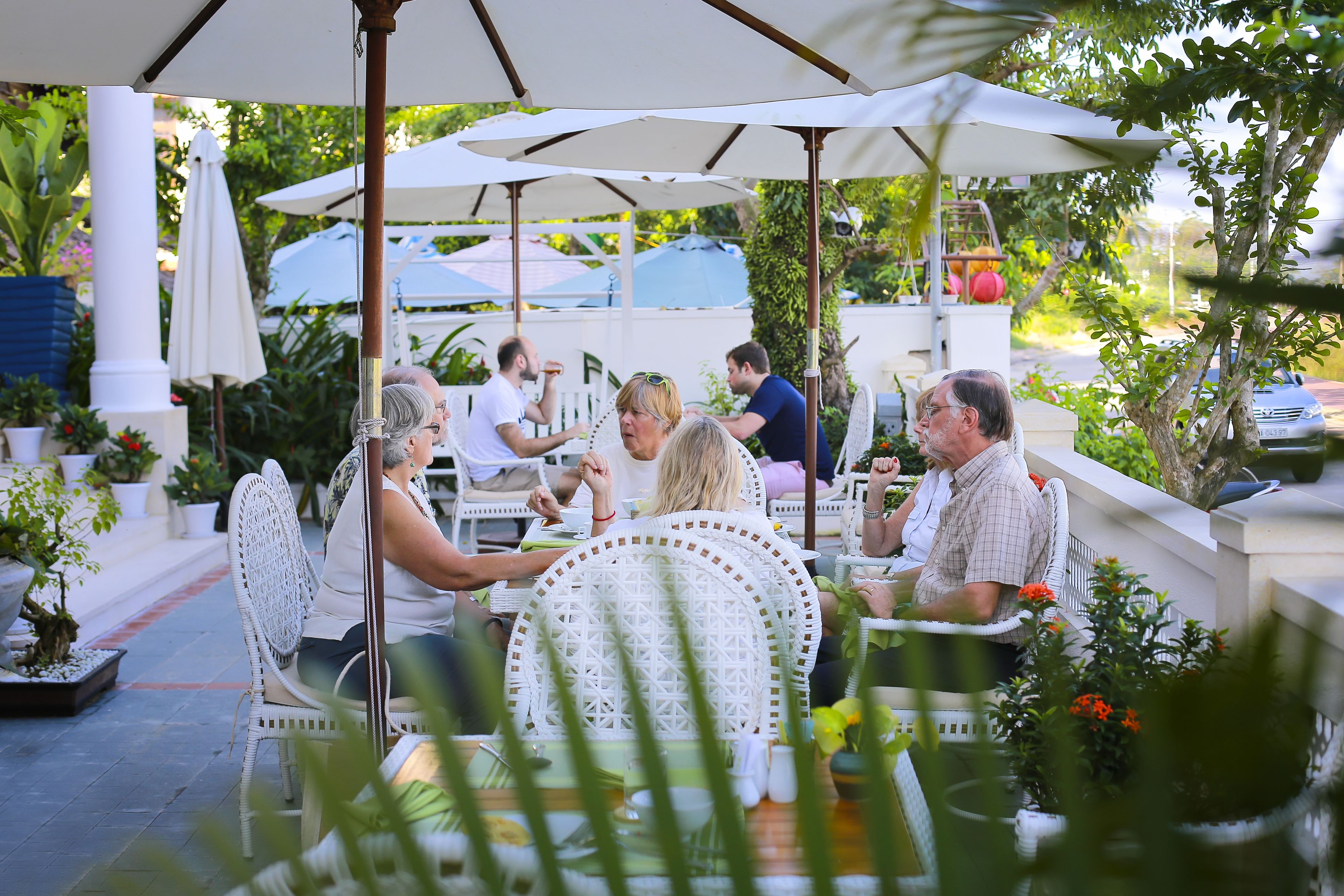 A group of people seated at tables under umbrellas in an outdoor setting with potted plants and white furniture.