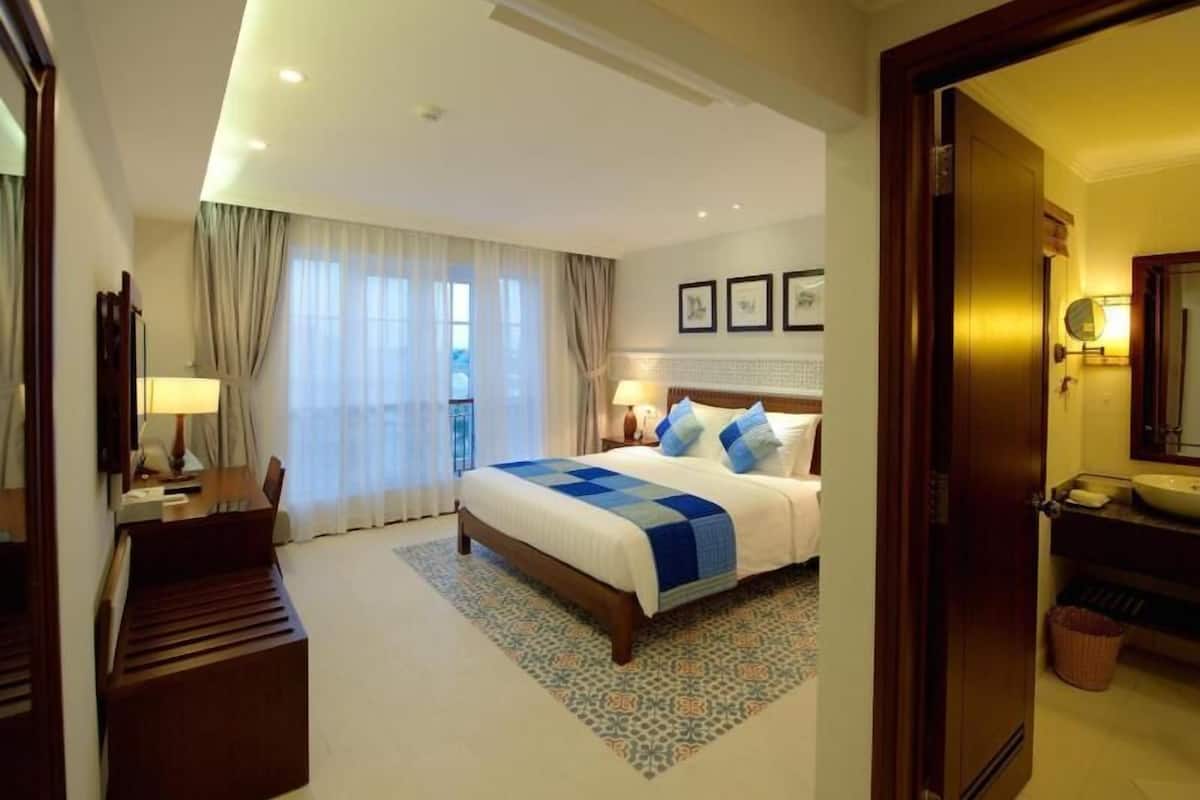 Habitación de hotel con una cama grande, un escritorio, una silla, un baño con lavamanos y espejo, y vistas al exterior.