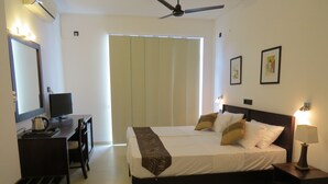 A hotel room with a bed, bedside table, lamp, desk with a computer and kettle, and a ceiling fan. - Chapelton House (Colombo)