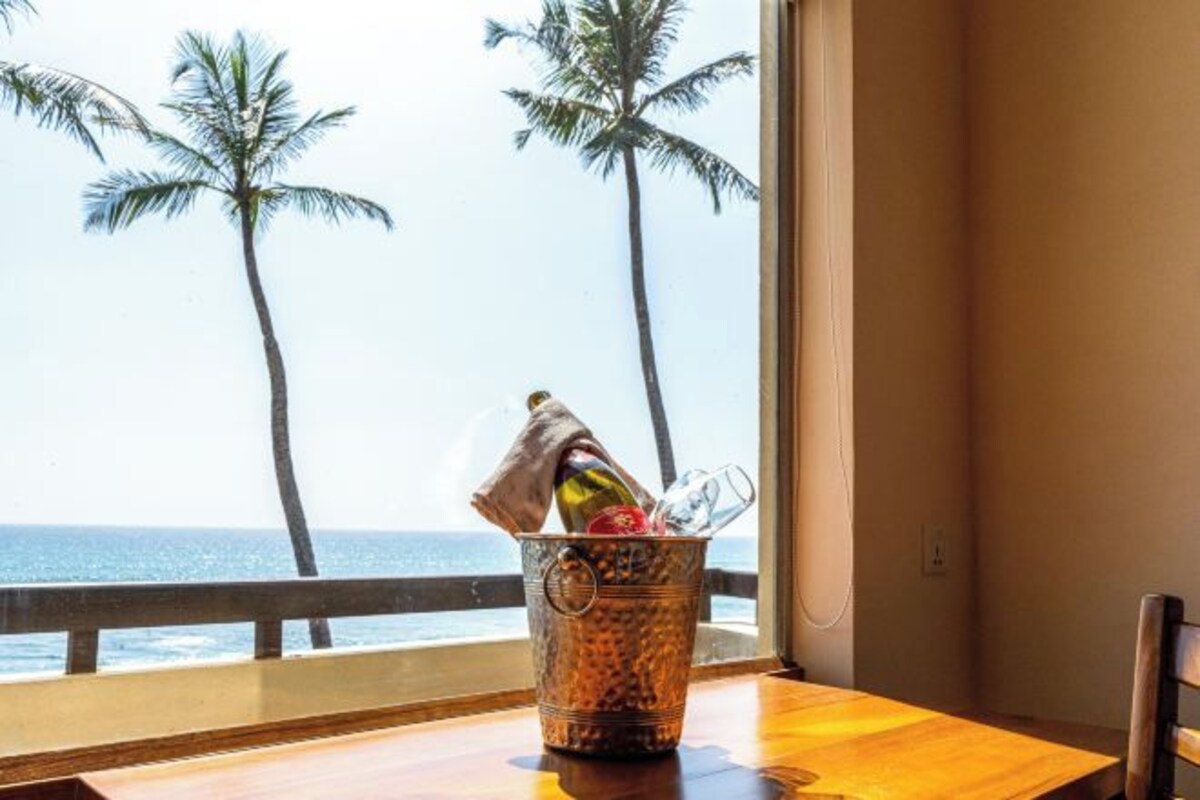 A metal bucket with a bottle and glasses on a wooden table by a window with a view of the ocean and palm trees.