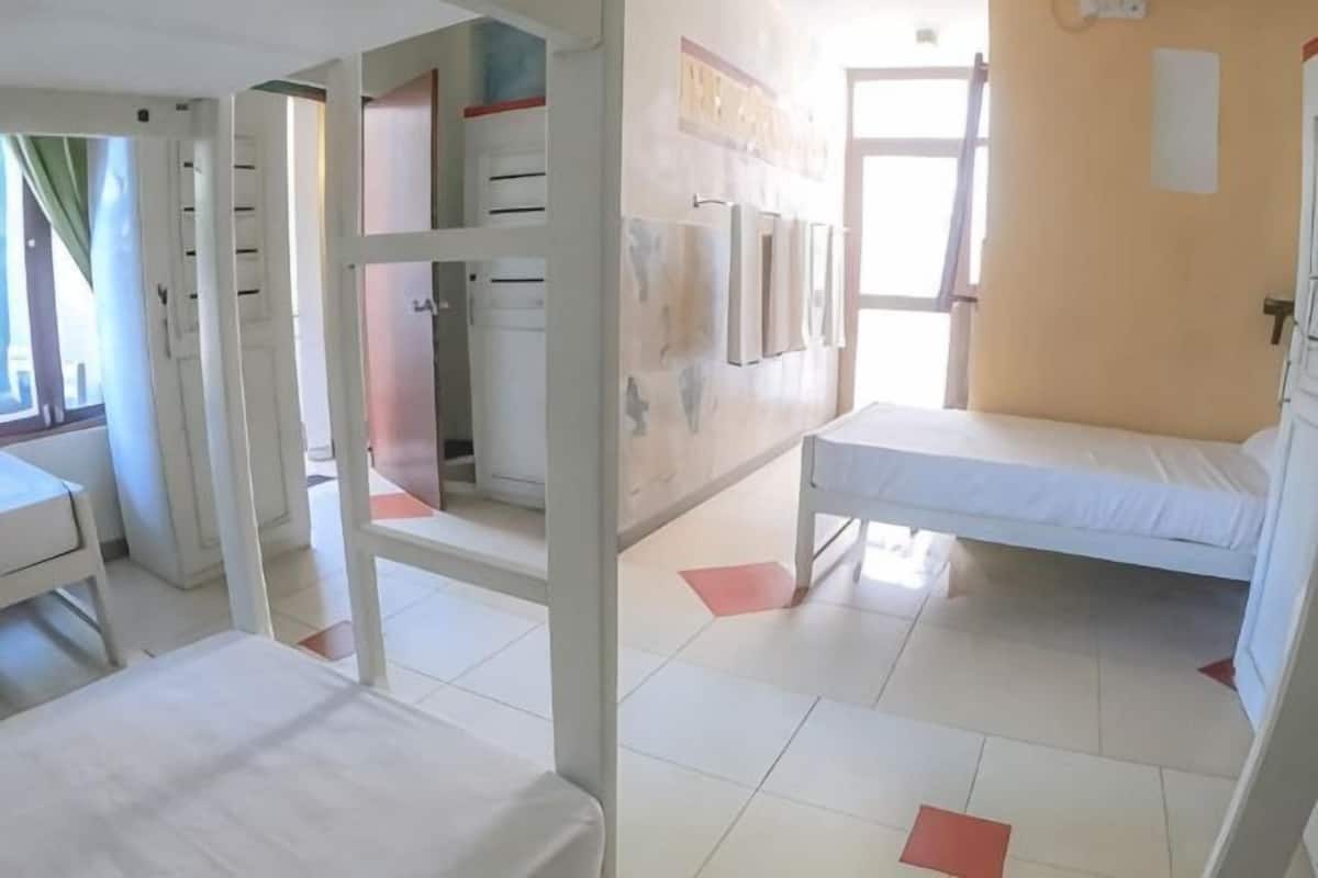 A room with a bed, a desk, and a chair. There is a window with curtains, a door, and a wall-mounted light fixture.