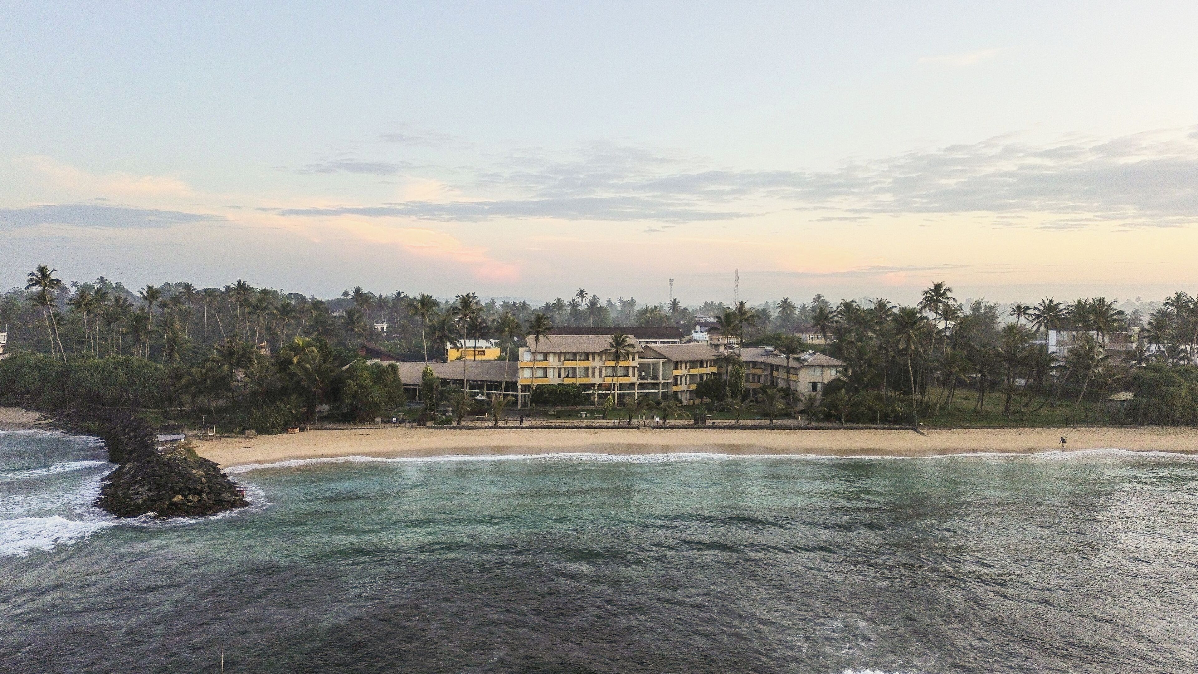 Un hotel frente a la playa con varios pisos, una playa de arena y palmeras al fondo.
