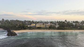 Un hotel frente a la playa con varios pisos, una playa de arena y palmeras al fondo.