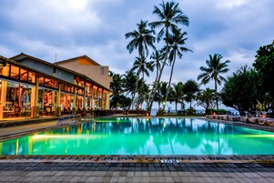 A swimming pool with a clear blue water surface, surrounded by palm trees and a building with large windows and outdoor seating.