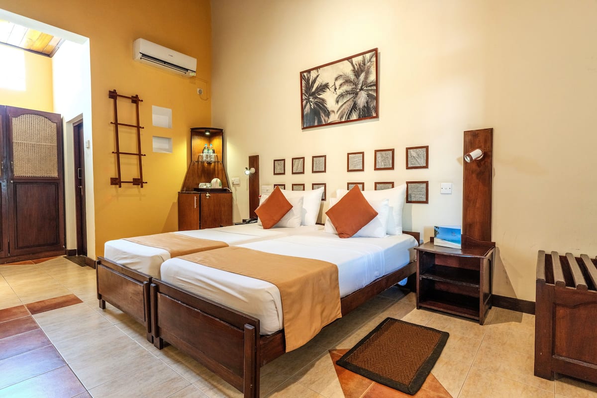 A hotel room with two beds, a wooden headboard, a wall-mounted air conditioner, a framed picture, and a small wooden nightstand.
