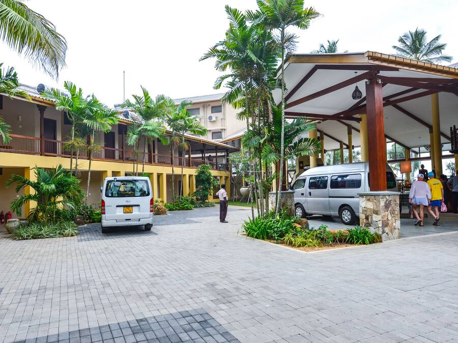 Entrada de hotel con camino de entrada pavimentado, vehículos estacionados y un área de estar al aire libre cubierta con palmeras.