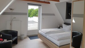A bedroom with a sloped ceiling, a bed with white bedding, a black chair, a radiator, and a window with a view of greenery. - Pension Marktblick (Friedrichstadt)