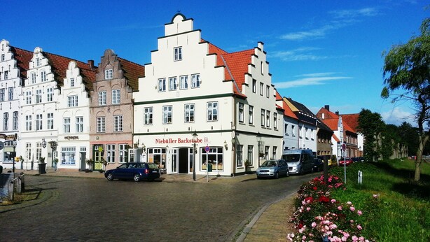 A cobblestone street lined with historic buildings, a sign for 'Niebüller Backstube', and parked cars. - Pension Marktblick (Friedrichstadt)