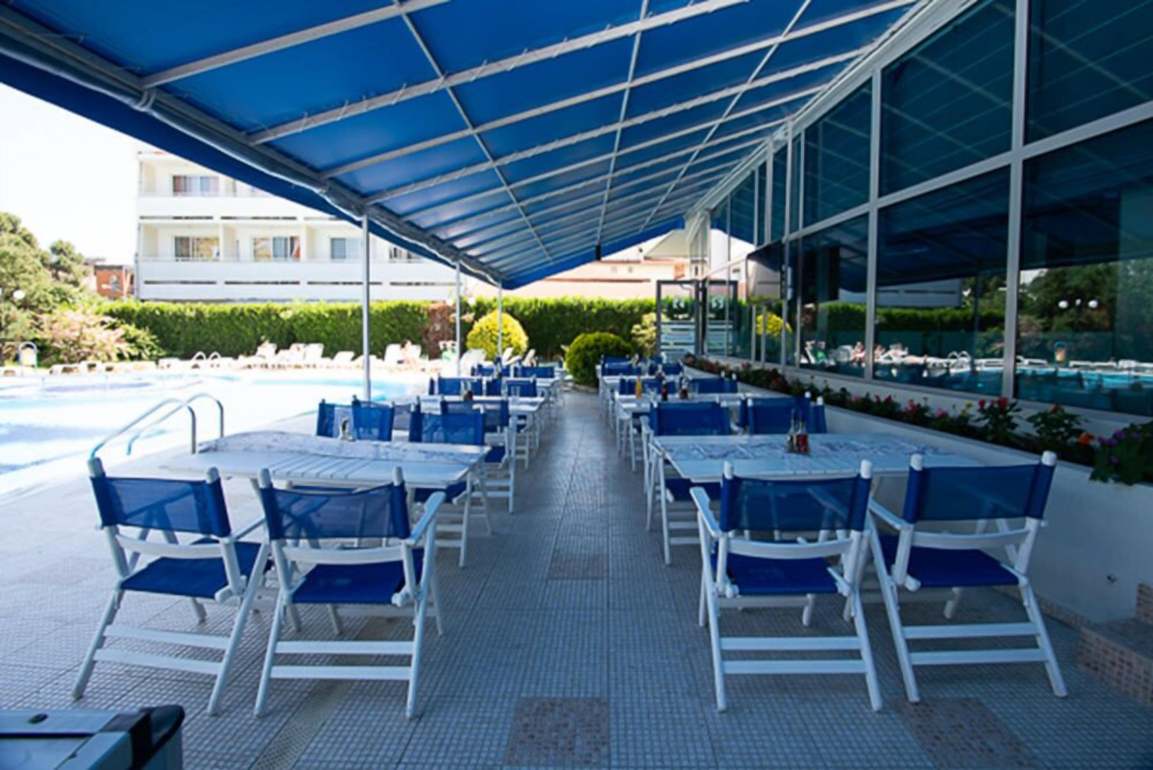An outdoor pool area with blue chairs and tables under a blue canopy, surrounded by a pool and a building with large windows.