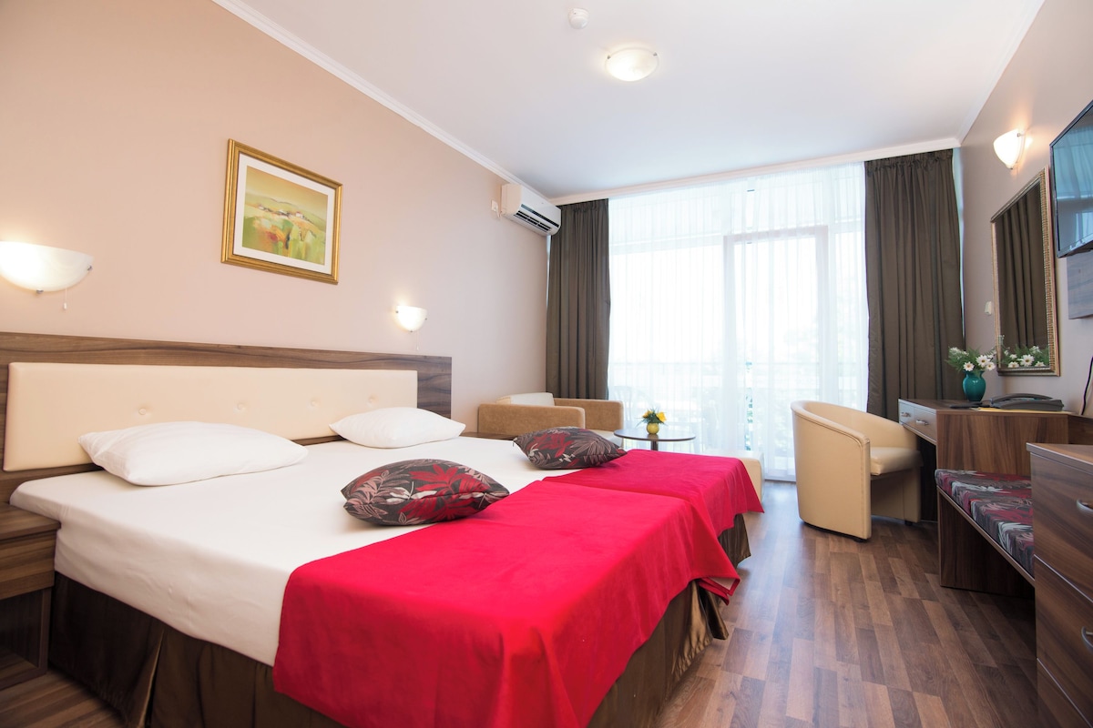Ein Hotelzimmer mit zwei Betten, einem Schreibtisch, einem Stuhl, einem Fernseher und einem großen Fenster mit Vorhängen.