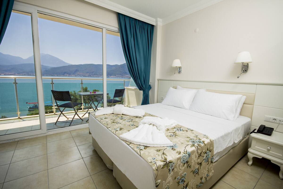 A hotel room with a bed, a chair, a nightstand, a lamp, and a view of the sea.