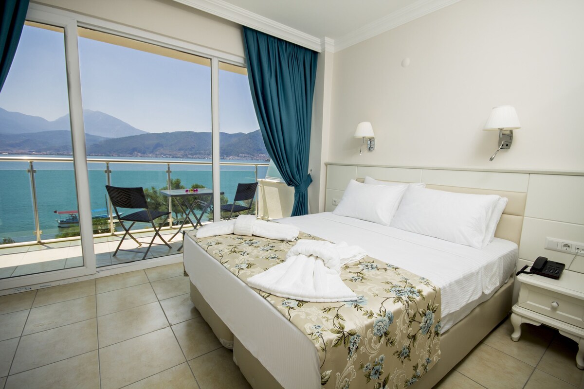 A hotel room with a bed, a chair, a nightstand, a lamp, and a view of the sea.