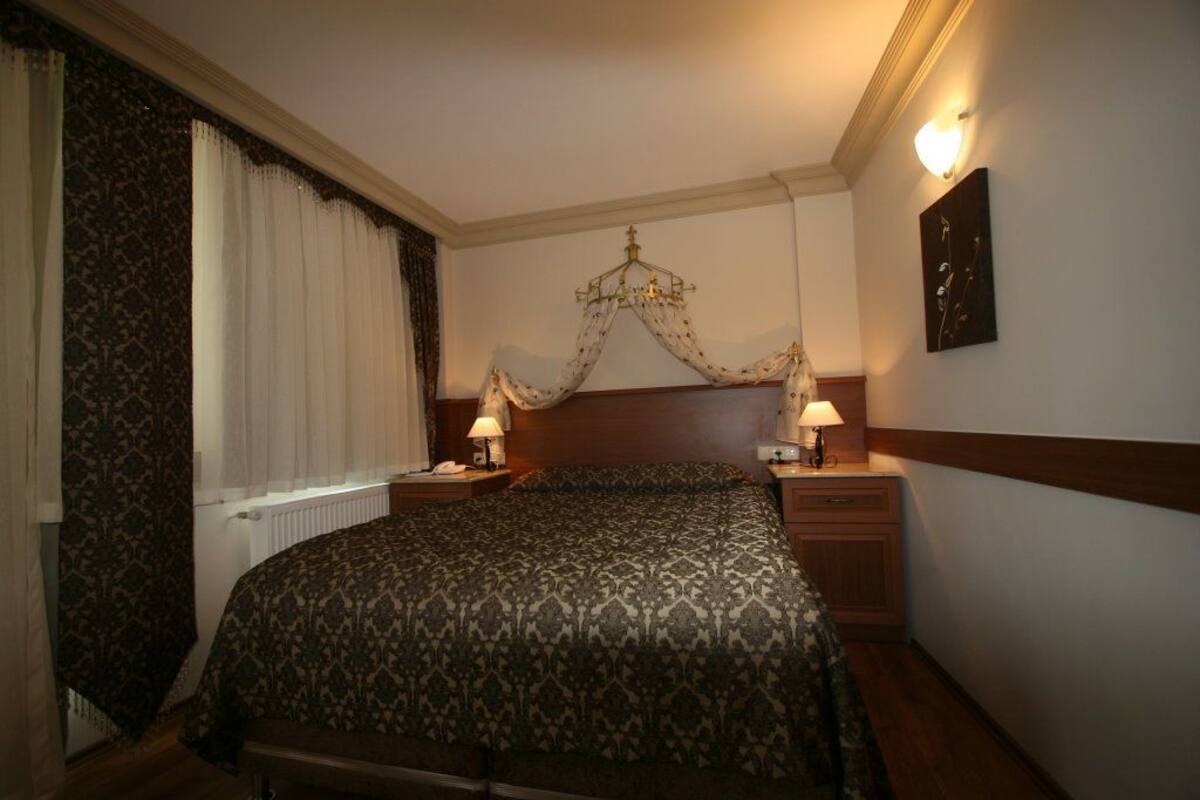 Une chambre d’hôtel avec un grand lit, deux tables de chevet avec des lampes, un lustre et une fenêtre avec des rideaux.