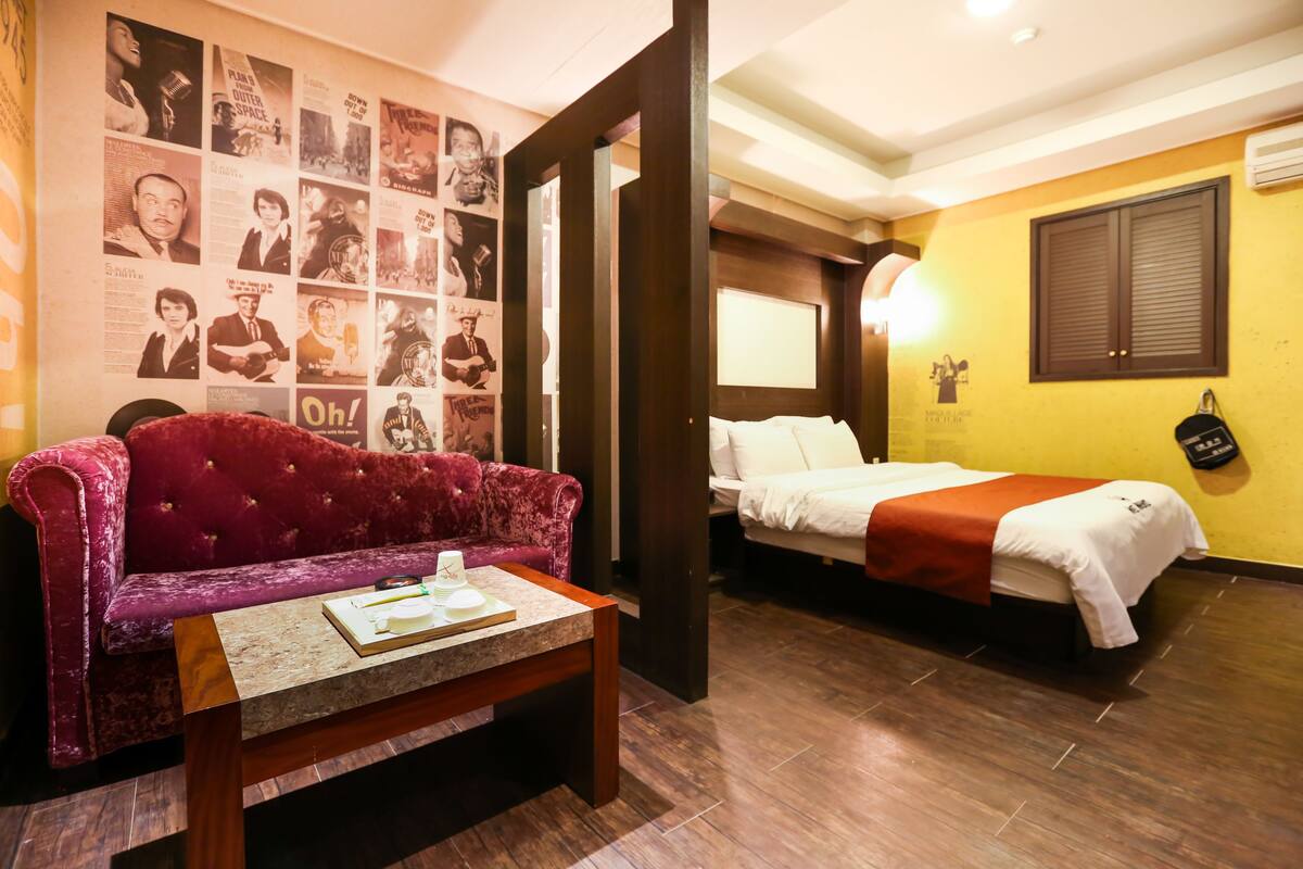A hotel room with a red velvet sofa, a wooden coffee table, a white bed with a red blanket, and a wall decorated with black and white photos.