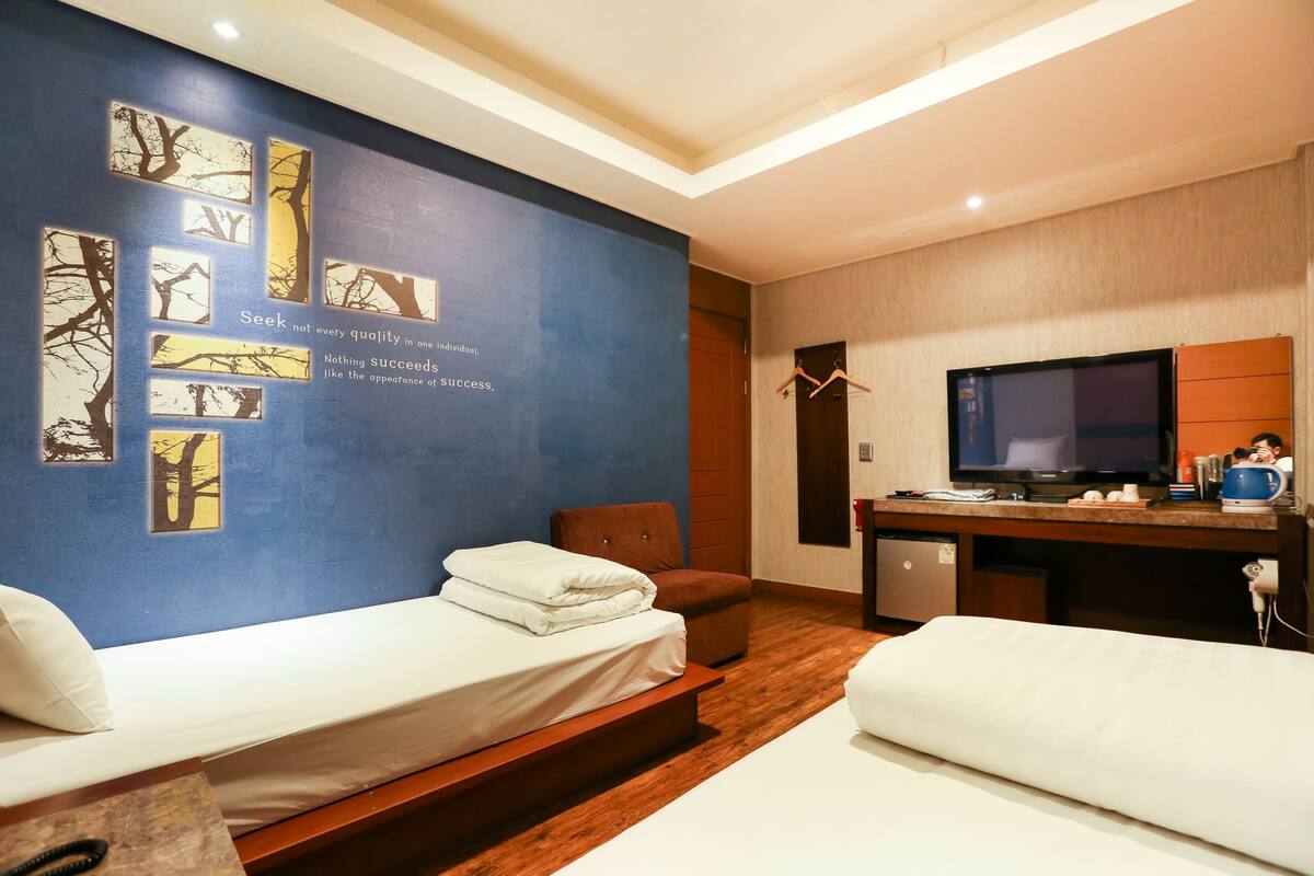 A hotel room with two single beds, a desk with a television, and a wall with tree artwork.