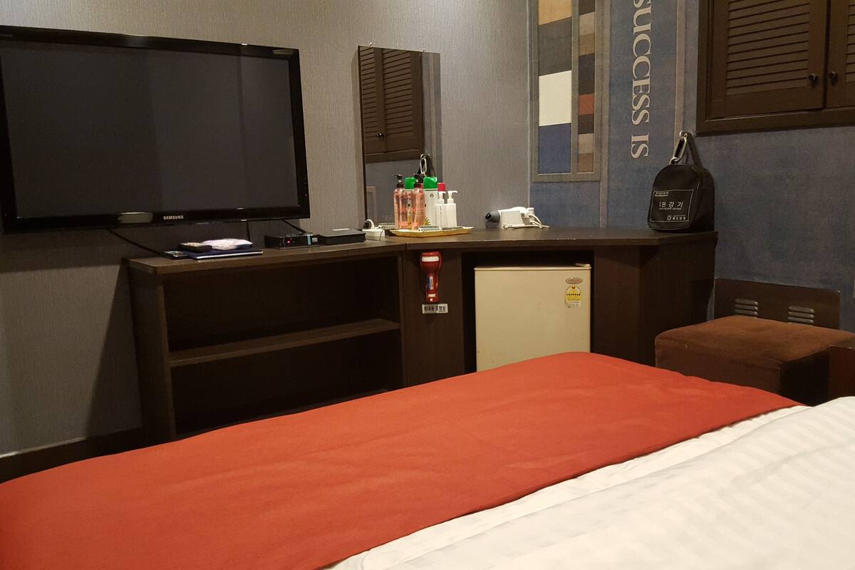 A hotel room with a bed, a TV, a desk with a computer, a mini-fridge, and a small table with toiletries.