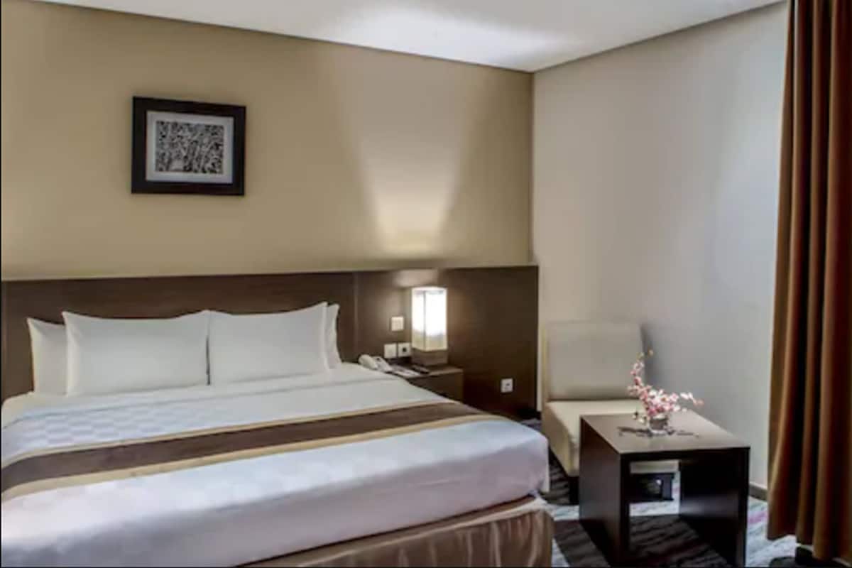 A hotel room with a large bed, a bedside table with a lamp, a chair, and a framed picture on the wall.
