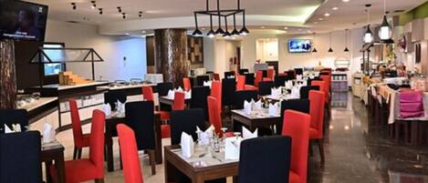 Un restaurante con sillas rojas y negras, mesas dispuestas con manteles blancos y una lámpara de araña moderna.