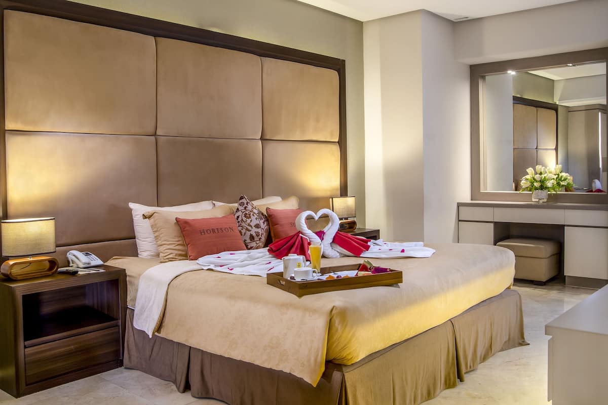 A hotel room with a large bed, a bedside table with a lamp, a telephone, and a tray with breakfast items.