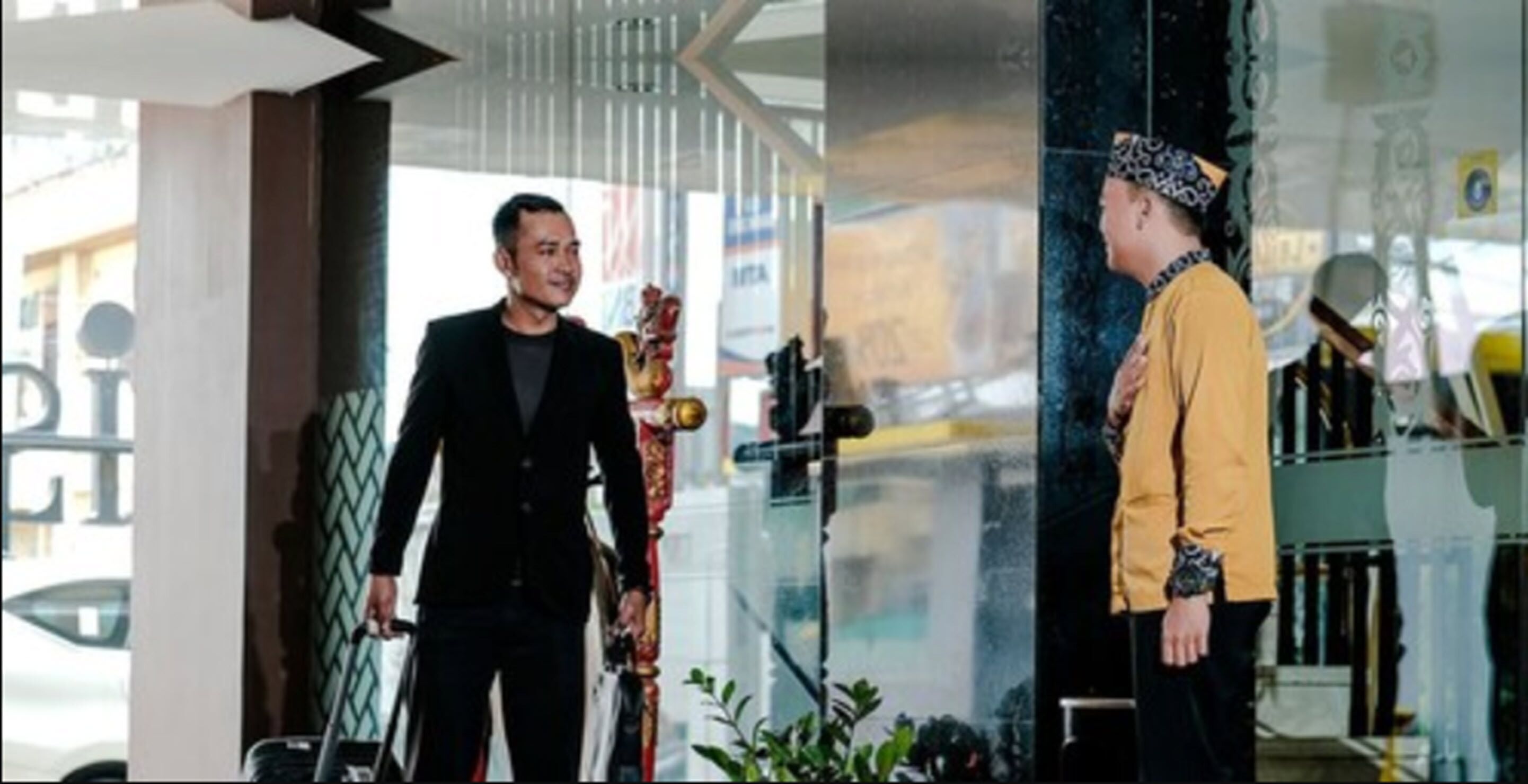 A man in a black suit holding a trophy, standing in a modern lobby with glass walls and a staircase.