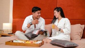 A couple sitting on a bed with a magazine, a tray of snacks, and a lamp on a bedside table.