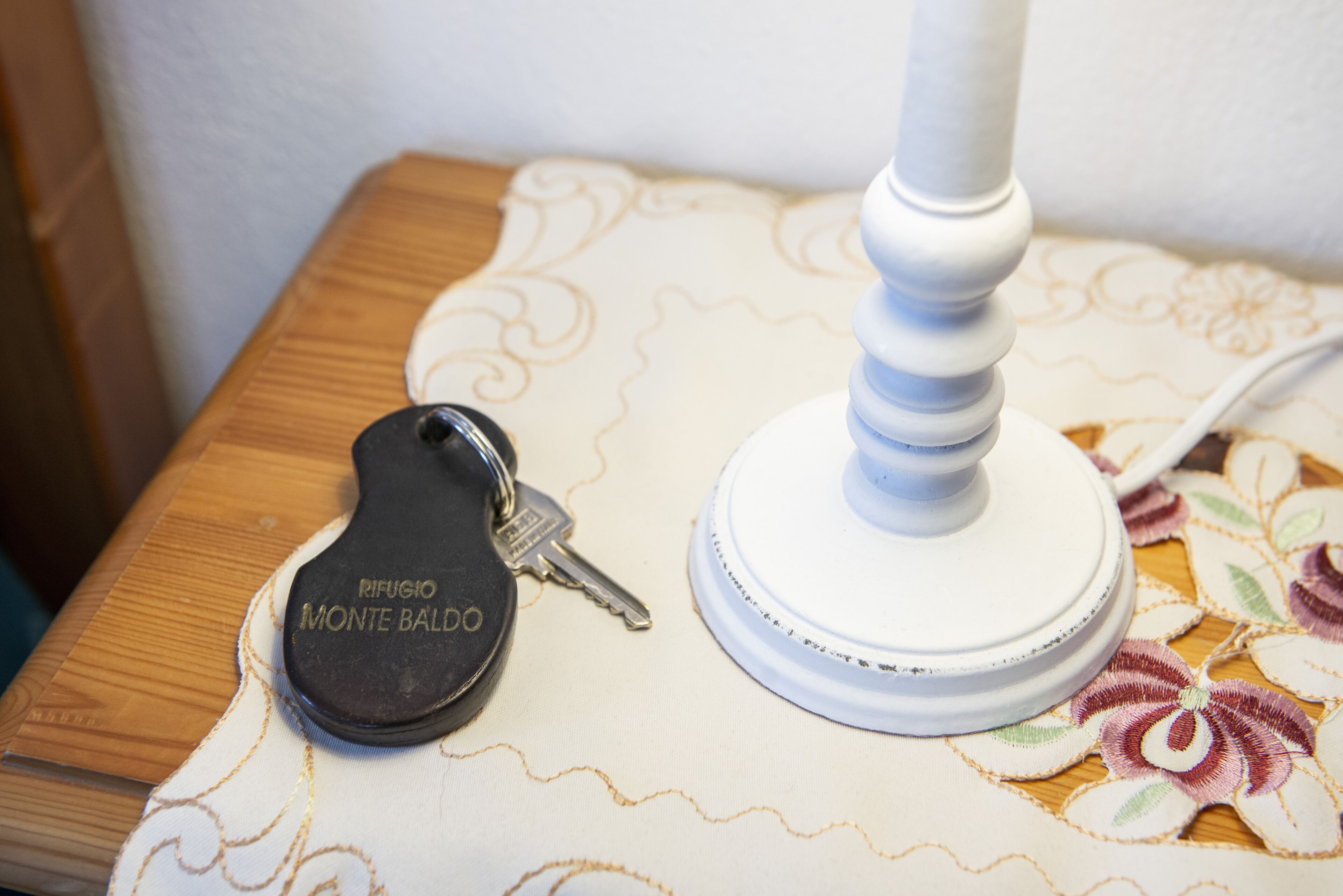 A wooden nightstand with a white lamp and a black keychain labeled "Rifugio Monte Baldo" on a floral-patterned tablecloth.