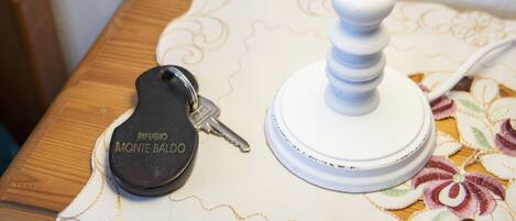 A wooden nightstand with a white lamp and a black keychain labeled "Rifugio Monte Baldo" on a floral-patterned tablecloth.