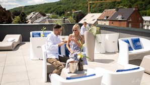 Una terraza en la azotea con sillones blancos, una pareja sentada a una mesa con bebidas, y una vista panorámica de casas y follaje al fondo.
