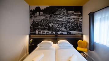 Habitación de hotel con dos camas, una silla amarilla, una fotografía grande en blanco y negro en la pared y una ventana con cortinas.