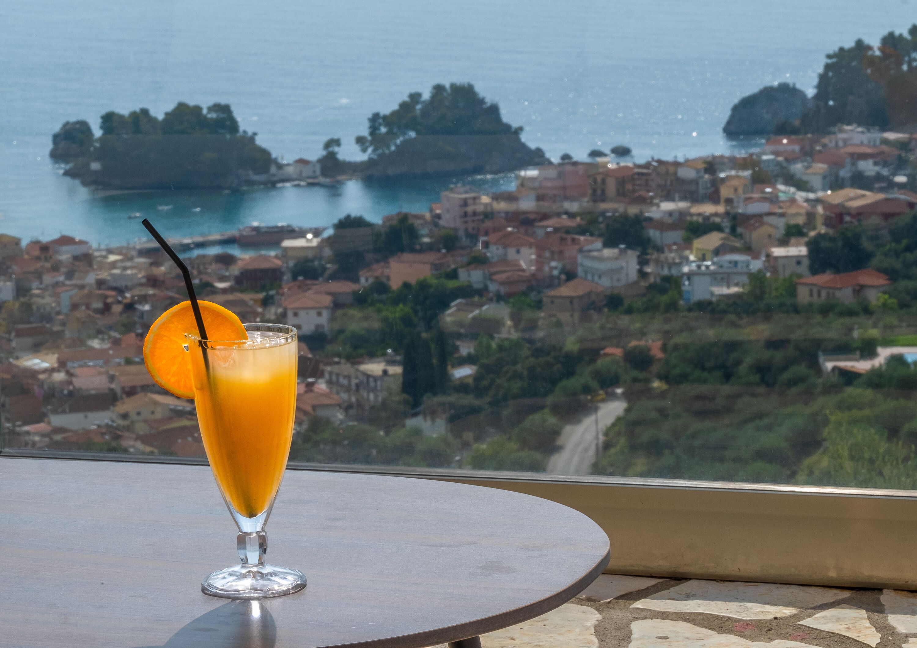 A glass of orange juice with a straw and a slice of orange on a table with a view of a coastal town.