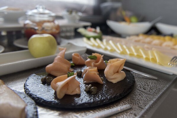 A variety of food items on a buffet table, including a plate with sliced vegetables, a dish with smoked salmon, and a plate with pasta.