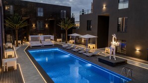A rectangular swimming pool with lounge chairs and umbrellas, surrounded by modern buildings with balconies and outdoor lighting. - Nacala Plaza Business Design Hotel (Nacala)