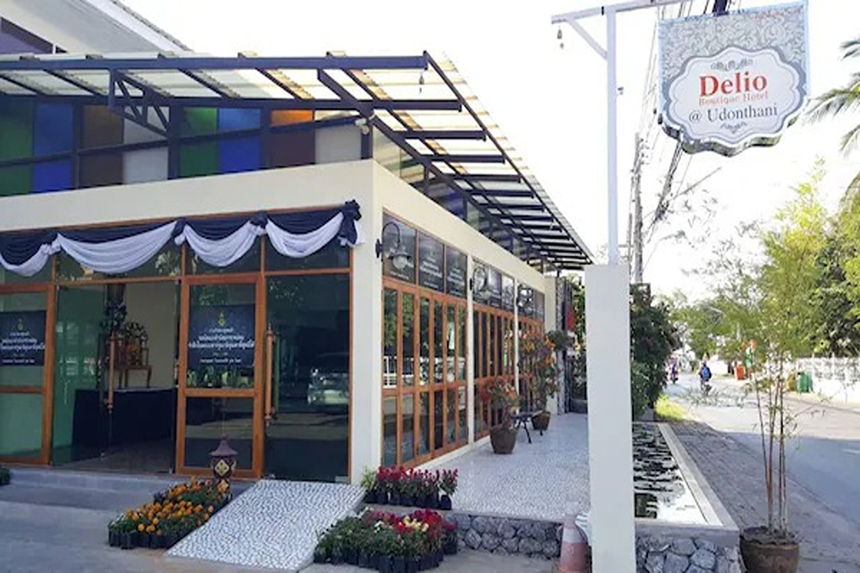 Un hôtel doté d’un espace extérieur couvert avec des sièges, de grandes fenêtres et d’une enseigne indiquant « Delio Bestiguer Hotel @ Udonthani ».