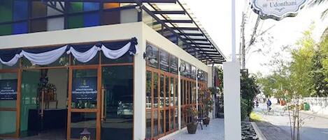Un hôtel doté d’un espace extérieur couvert avec des sièges, de grandes fenêtres et d’une enseigne indiquant « Delio Bestiguer Hotel @ Udonthani ».