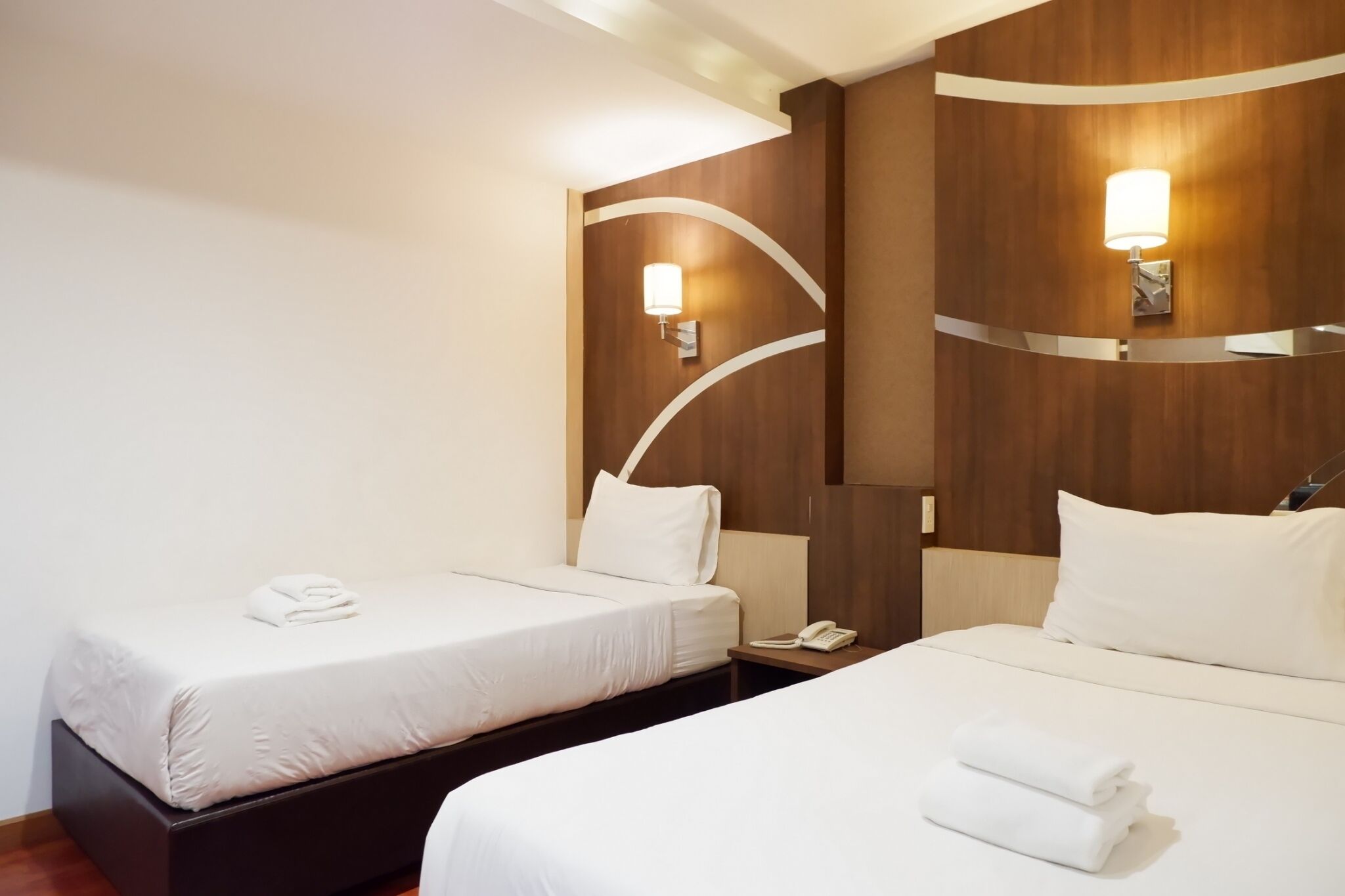 Une chambre d’hôtel avec deux lits, une tête de lit en bois, une table de chevet avec un téléphone et des lampes fixées au mur.