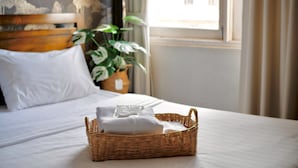 Un lit bien fait, avec du linge de lit blanc, un panier en osier contenant des serviettes pliées et, au fond, une plante en pot.