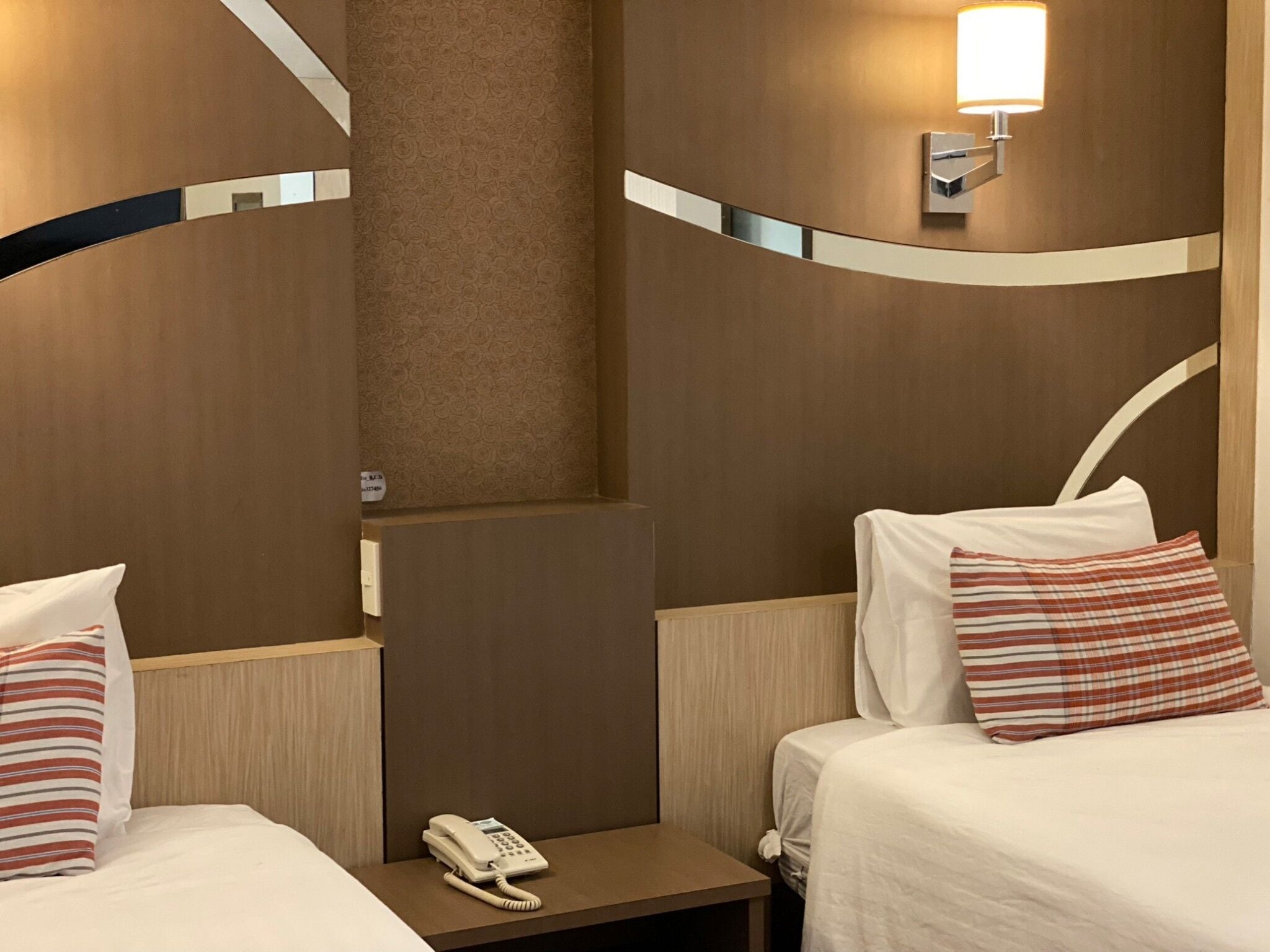 A hotel room with two beds, a bedside table with a phone, and a wall-mounted light fixture.