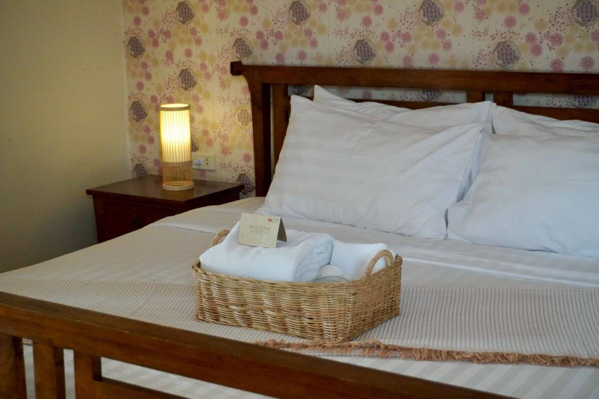 A neatly made bed with white linens and pillows, a wicker basket with folded towels, a lit bedside lamp, and a welcome sign on the nightstand.