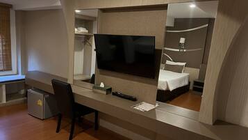 A modern hotel room with a large TV, a desk with a chair, a small fridge, and a bed with a white bedspread.